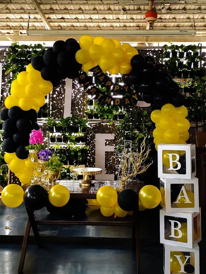 A modern and stylish balloon arch in a striking yellow and black color scheme. Set against a natural green wall, this setup includes a 'Happy Birthday' banner and transparent 'BABY' blocks for a chic, contemporary feel.