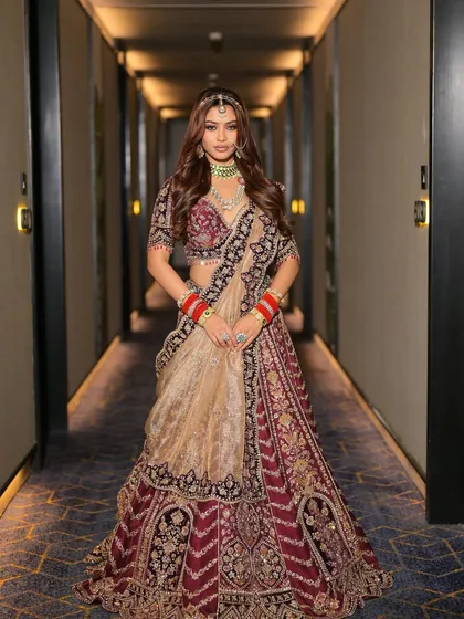 A full-length shot in a modern setting, showcasing a contemporary bridal look. The makeup is designed to complement the rich colors of the lehenga and the bride's confident stance.