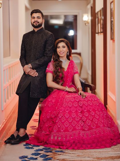 A regal portrait of the couple at their reception. The bride is seated while the groom stands beside her, their vibrant outfits creating a striking visual.