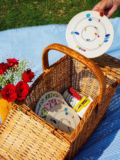 Another angle of the picnic scene, showing the basket being filled with artisanal tableware. The sunny, outdoor setting is very appealing.