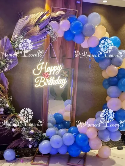 A modern and chic circular photo frame decorated with blue and beige balloons and dried pampas grass, with a "Happy Birthday" neon sign in the center.