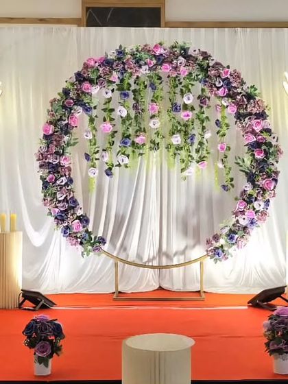 A breathtaking wedding stage with a circular arch adorned with hanging purple flowers. The large flower lamps and red carpet create a dramatic and luxurious feel.