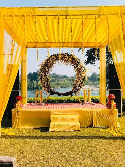 A beautiful outdoor Haldi setup by the water. The simple yellow mandap with a large floral wreath creates a stunning focal point against the scenic backdrop.