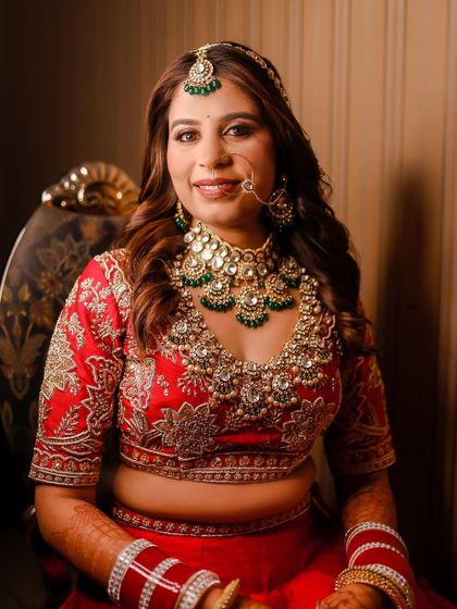 A radiant bridal portrait. The bride is seated, smiling warmly at the camera, showcasing her elaborate green and white stone necklace and the detailed embroidery on her red blouse.