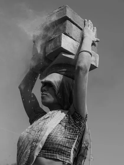 A black and white portrait of a woman lifting a heavy load of bricks, her determined profile and the dust in the air emphasizing her strength.