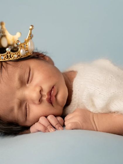 A close-up portrait of a sleeping newborn wearing a detailed gold and pearl crown, showcasing their peaceful expression.