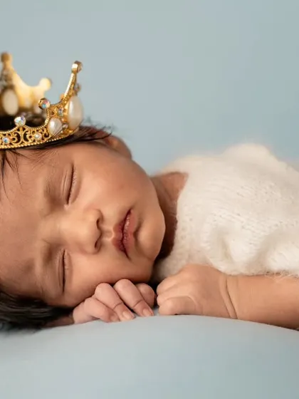 A close-up portrait of a sleeping newborn wearing a detailed gold and pearl crown, showcasing their peaceful expression.
