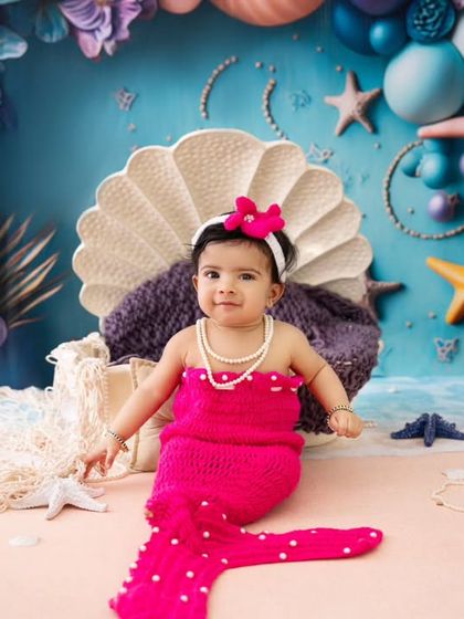 A sweet little mermaid has arrived. This six-month-old is all smiles in her bright pink mermaid outfit, sitting in a clamshell throne against a beautiful ocean-themed backdrop.