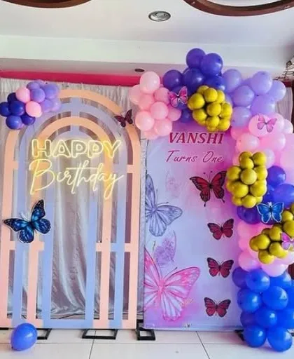 A butterfly-themed first birthday party with a unique wooden arch backdrop. The setup includes a "Happy Birthday" neon sign and a colorful balloon arrangement.