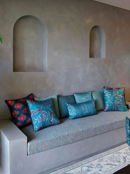 A built-in sofa against a grey lime-washed wall with arched niches. The space is brought to life with vibrant teal and floral cushions, adding a pop of color and personality.
