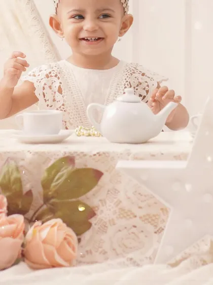 A delightful tea party themed shoot, featuring a smiling baby girl enjoying a pretend cup of tea at a beautifully set lace table.