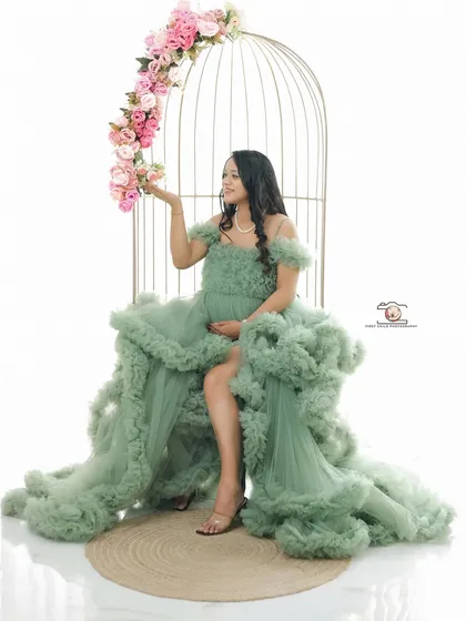 A whimsical and creative studio portrait. The mother-to-be is seated in a birdcage-like prop, adorned with flowers, wearing a beautiful ruffled green gown that makes her look like a fairy-tale character.