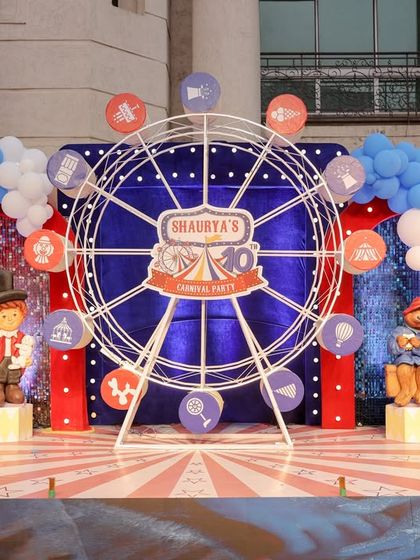 This stunning carnival party backdrop features a large, illuminated ferris wheel as the centerpiece, surrounded by elaborate balloon arrangements and charming character figures.