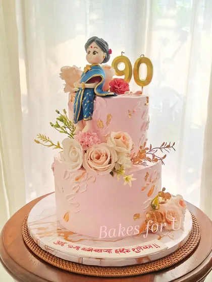 A truly special two-tier cake for a 90th birthday, featuring a handcrafted fondant figure of the matriarch in a beautiful blue saree. The cake is written in Marathi, adding a personal, local touch.