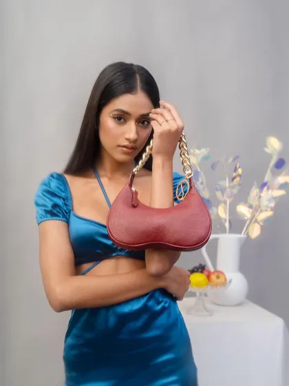 A model poses with a red Sarjaa handbag in a studio setting. The composition draws the eye directly to the product, highlighting its unique shape and gold chain detail.