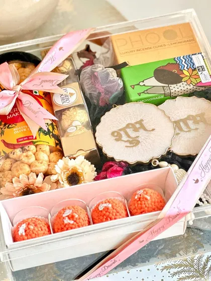 We believe a gift should be an experience. This clear acrylic box for Diwali is filled with festive joy, including traditional ladoo-shaped candles, Shubh Labh wall hangings for blessings, gourmet makhana, and rich chocolates, all tied together with a personalized ribbon.