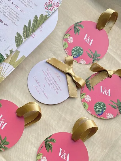 Bright pink, circular welcome tags for guest hampers. The design features a peacock and tropical flowers, tied with a gold ribbon for a touch of luxury.