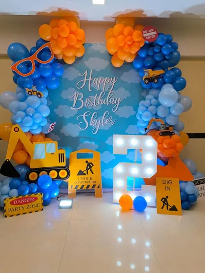A construction themed second birthday party. The setup includes a blue and orange balloon arch, cutouts of excavators and trucks, and fun props like 'Under Construction' signs.