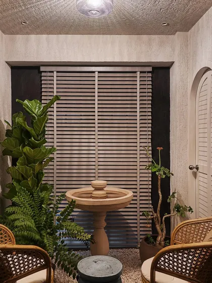 In the Zen House, I designed this tranquil fountain area as a meditative retreat. A custom grey stone fountain sits on a bed of white gravel, surrounded by plants and cane chairs, creating a serene indoor garden.