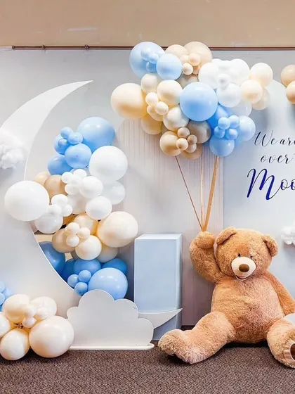 An adorable "We are over the Moon" teddy bear theme, with a large moon prop and cloud cutouts surrounded by blue, white, and beige balloons.