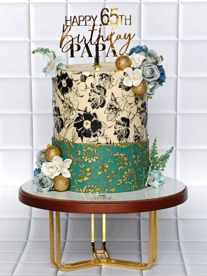 This two-tiered cake was designed for a 65th birthday, featuring a hand-stenciled floral pattern on the top tier and a gold-embossed damask design on the bottom. The soft color palette and floral accents make it a distinguished and personal centerpiece.