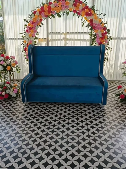 A simple and elegant floral arch for a small event or photo booth. This setup features a blue sofa and two floral stands, perfect for a daytime function.