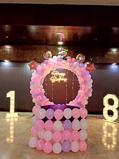 A pink and purple themed 18th birthday party in a banquet hall. The decoration includes a balloon ring with a neon sign and large light-up numbers.