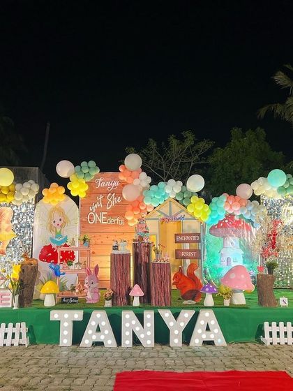 A complete outdoor setup for an "Enchanted Forest" first birthday party, featuring a large stage with a wooden backdrop, mushroom props, character cutouts, and a light-up name sign.