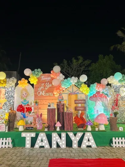 A complete outdoor setup for an "Enchanted Forest" first birthday party, featuring a large stage with a wooden backdrop, mushroom props, character cutouts, and a light-up name sign.