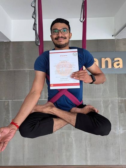 A male student demonstrates a seated lotus pose in the air, proudly displaying his aerial yoga certification.