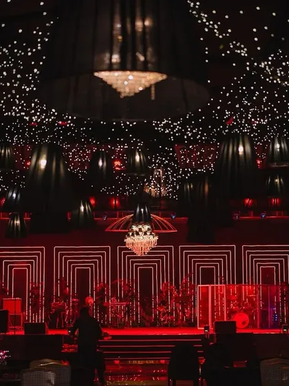 A dazzling setup for a sangeet night, where a ceiling of countless lights and hanging lamps creates a celestial glow. The stage, lit in dramatic red with geometric patterns, sets the scene for an unforgettable evening of performances and celebration.
