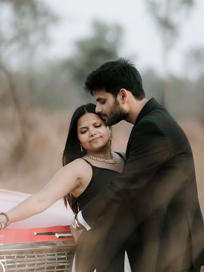 A tender moment with the groom kissing the bride's forehead as they lean against a vintage red car, creating a soft and romantic image.