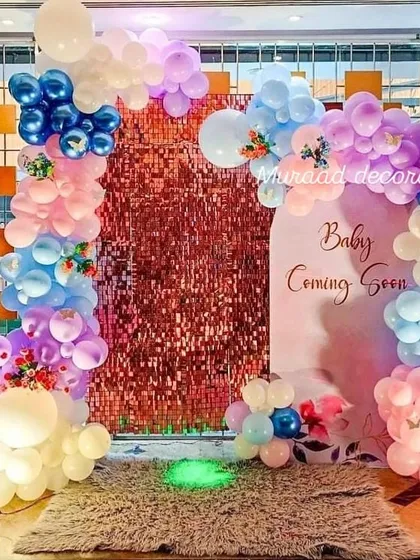 A vibrant 'Baby Coming Soon' baby shower setup. This decoration combines a shimmering rose gold backdrop with a colorful balloon arch in pink, blue, and purple, suitable for a celebration that isn't revealing the gender.