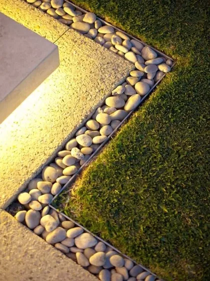 A close-up detail showing a clean border between a lawn and a paved area, filled with smooth river pebbles and accented with lighting. This is a modern and stylish finishing touch.