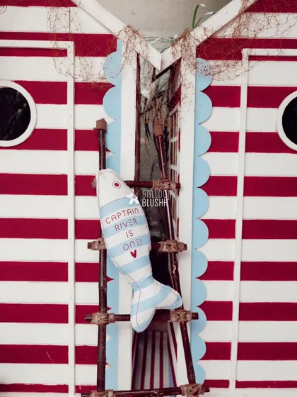 A custom-made fish-shaped pillow with the words 'Captain River is One!' hangs on a rustic ladder against the striped sailor cabin.