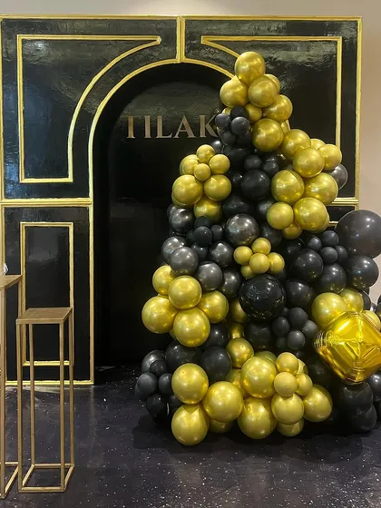 A classic and bold black-and-gold theme for a 50th milestone birthday. The decor features a grand arch backdrop with the name 'Tilak' and a cascading organic balloon arrangement.