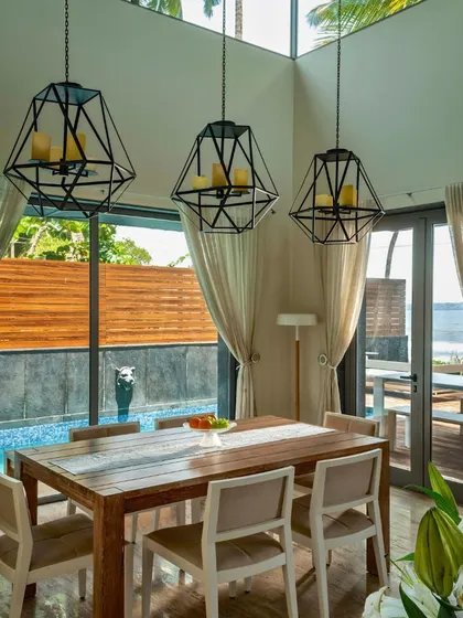 A dining room in a Goa villa, featuring geometric pendant lights over a rustic wooden table. The double-height space and large windows connect the dining experience directly to the poolside view and coastal landscape.
