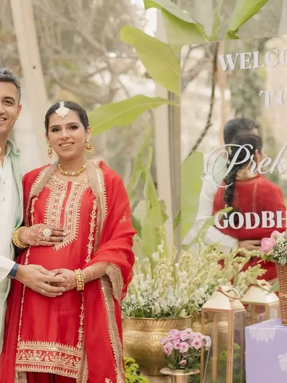The happy couple at their Godh Bharai ceremony. The mom-to-be is radiant in her traditional red attire, standing beside the custom acrylic welcome sign.