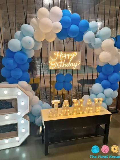 A close-up of a stylish blue and white themed birthday decoration, featuring a circular balloon arch, a neon sign, and light-up letters for the name and age.