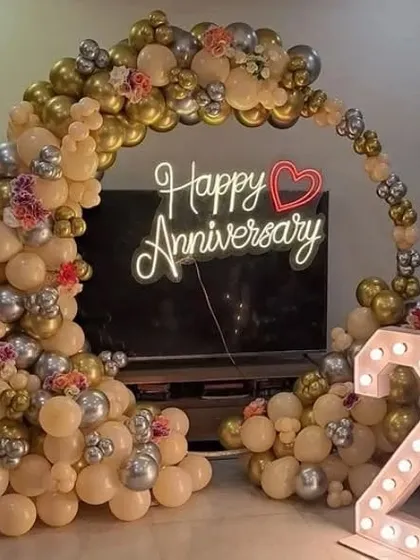 An anniversary celebration with a balloon arch in peach, gold, and silver. The setup includes a "Happy Anniversary" neon sign and marquee numbers.