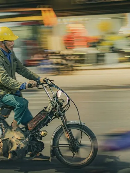 A motion-blur street photograph from Hanoi, Vietnam, capturing a man in a yellow helmet riding a vintage moped, creating a sense of movement against a blurred urban background.