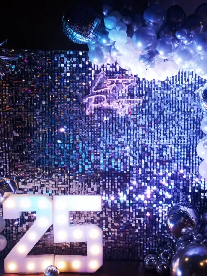 Celebrating a 25th anniversary with a touch of disco fever. This setup includes a silver sequin wall, a "Let's Party" neon sign, and a black and silver balloon garland with starburst accents.