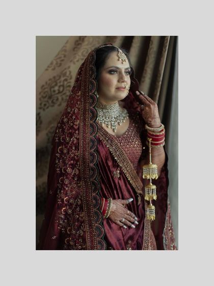 A classic bridal portrait. The bride looks elegant and poised in her deep red lehenga, captured in a moment of quiet reflection by the window.