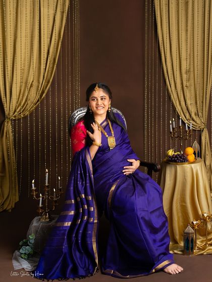 A joyful portrait of an expecting mother in a vibrant blue silk saree. She is seated on a throne-like chair in a studio decorated with golden curtains and traditional elements.