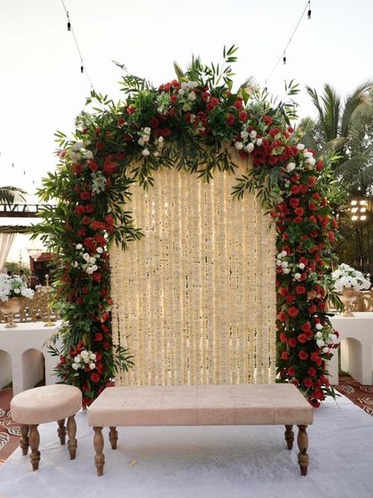 An intimate photo booth or seating area for a Nikah ceremony. The arch is decorated with red and white roses and features a backdrop of delicate white flower strings, creating a romantic and detailed corner.