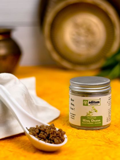 This shot of Hing Daana is set in a traditional kitchen scene with brass utensils and a white cloth. The warm lighting and rustic props evoke a sense of authentic, home-cooked meals.