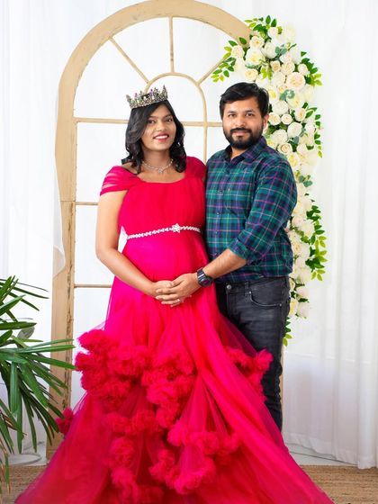 A happy couple's portrait in our studio. The mom-to-be wears a vibrant pink gown and crown, sharing a joyful moment with her partner against our floral archway backdrop.