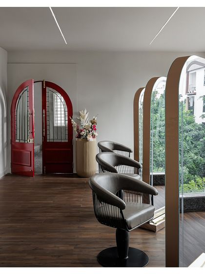The styling stations feature custom-arched mirrors and comfortable, modern chairs. A striking red arched door adds a bold pop of color and architectural interest, while large windows provide ample natural light.