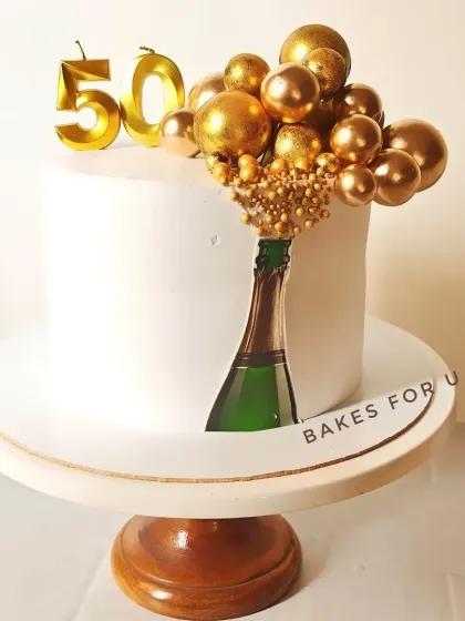 A chic champagne-themed cake for a 50th birthday. This all-white cake features an edible image of a champagne bottle and a cascade of golden chocolate spheres.