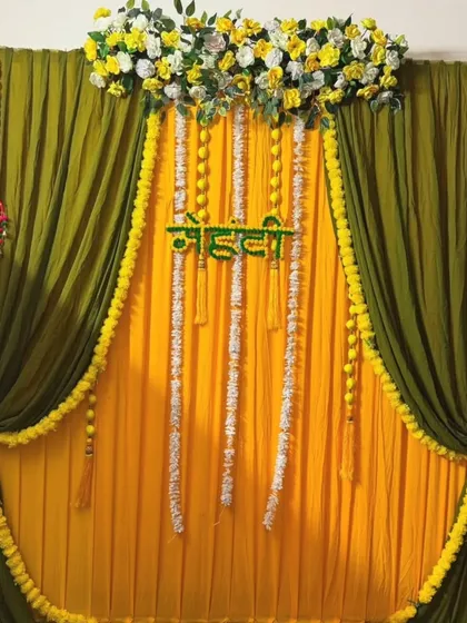 A simple yet beautiful Mehandi backdrop with layered green and yellow drapes. The decor is accented with strings of white and yellow flowers, creating a fresh and clean look for a home event.